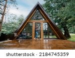 Cute barefoot woman stands on the terrace of triangular wooden cabin with a glass of herbal tea
