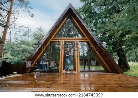 Cute barefoot woman stands on the terrace of triangular wooden cabin with a glass of herbal tea