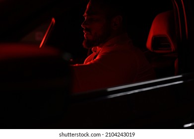 Cute Bearded Strong Young Man In White Shirt Sitting In Luxury Sport Muscle Car Lit With Red Light In Night City