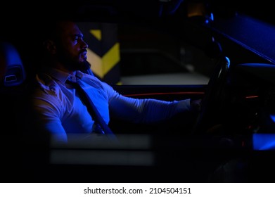 Cute Bearded Strong Young Man In White Shirt With Tie Sitting In Luxury Sport Muscle Car Lit With Blue Light In Underground Garage