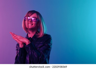 Cute beautiful blonde woman in leather jacket sparkly dress trendy sunglasses fold hands look at camera posing isolated in blue pink color light studio background  Neon party concept  Copy space