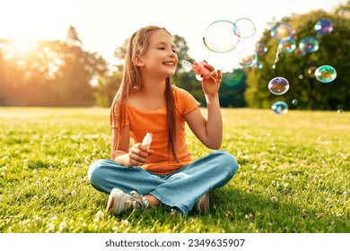 Cute beautiful girl blowing soap bubbles sitting on the grass in the meadow in the park, playing and having fun on a warm sunny weekend.