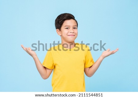 Cute bewildered mixed race boy in yellow t shirt looking at camera with smile and shrugging shoulders against blue background