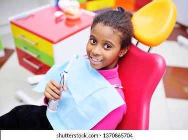 Cute Black Baby Girl Smiling Sitting In A Red Dental Chair At The Examination At The Children's Dentist