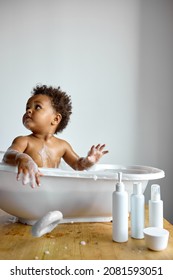 Cute Black Baby Girl Takes Bath, Looking Up. In White Bathroom. Girl Bathes In Basin Alone, At Home, In The Morning At Weekends. Healthy Boy Or Girl Playing, Splashing And Having Fun During Bathtime.