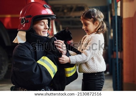 Cute black cat. Happy little girl is with female firefighter in protective uniform.