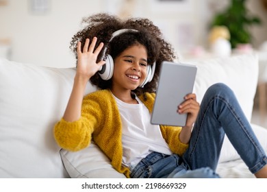 Cute Black Child Preteen Pretty Girl With Bushy Hair Having Online Party With Friends While Staying Home, Kid Sitting On Couch, Using Wireless Headset, Waving And Smiling At Digital Tablet Screen