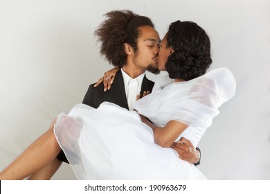 Cute black couple in romantic setting, she is wearing a white dress, and he is wearing a suit coat, he is holding her as they kiss