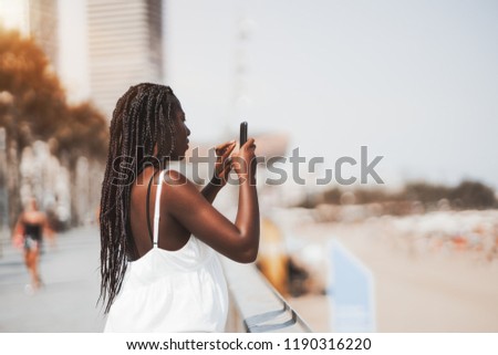 Cute black girl in a white dress is photographing sea and beach using the camera of her mobile phone; side view of a young African female with braids taking photos on the street via her smartphone