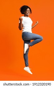 Cute black lady winner jumping up in the air and showing funny figures, having good day, showing positive emotions over orange background. African american woman posing at studio, having fun