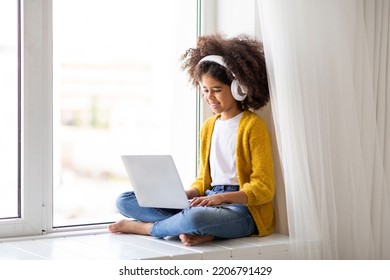 Cute Black Little Girl With Wireless Headphones Using Laptop At Home, Sitting On Window And Looking At Screen, Typing On Computer Keyboard, Playing Video Games On Computer, Copy Space