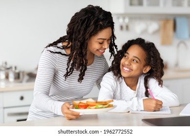 Cute Black Mother And Daughter Eating Sandwiches At Kitchen, Hugging And Smiling, Copy Space. Happy African American Mummy And Kid Having Snack, Painting Or Coloring Together At Home