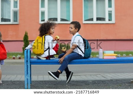 Cute Black schoolboy and girl eating outdoors next the school. Healthy school breakfast for child. Food for lunch, lunchboxes with sandwiches, fruits, vegetables, and water.
