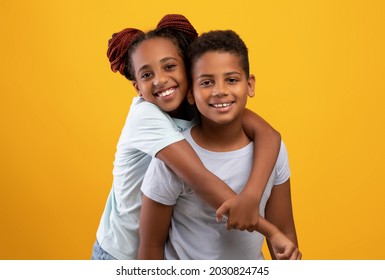 Cute Black Teen Girl Hugging Her Little Brother, Showing Her Love And Affection, Yellow Studio Background. Happy Loving Afro American Siblings In White T-shirts Embracing And Smiling At Camera