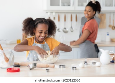 Cute Black Teen Girl Making Dough For Cookies While Her Beautiful Mother Washing Dishes, Kitchen Interior. Loivng African American Woman Mom Teaching Her Daughter Baking, Selective Focus On Girl