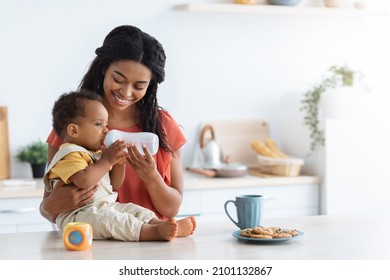 Cute Black Toddler Boy Drinking Water From Bottle While Spending Time With Smiling Young Mom In Kitchen, Thirsty Adorable Infant Baby Sitting On Counter Table And Enjoying Healthy Drink, Copy Space