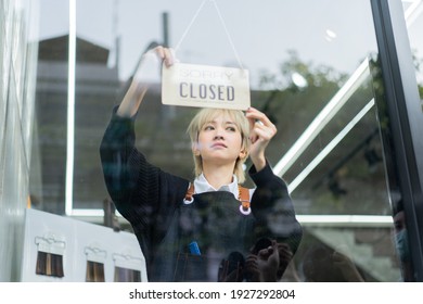 Cute blond Asian female hairdresser in navy cardigan and denim apron standing behind a glass door while turning SORRY CLOSED sign hanging on the glass door in hair salon due to lockdown measure.