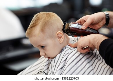 Cute blond baby boy with blue eyes in a barber shop having haircut by hairdresser. Hands of stylist with tools. Children fashion in salon. Indoors, dark background, copy space. - Powered by Shutterstock