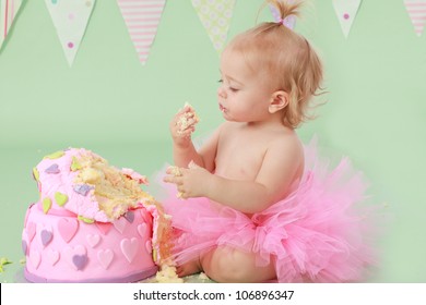 Cute blond baby girl in pink tutu and ponytail sitting on green background by  birthday party double tier pink and purple fondant iced cake with hands in cake happy and excited to celebrate and eat it