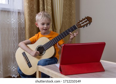Cute blond boy plays the guitar and looks at the tablet standing on the table. A fun online lesson on the internet. Digital education in self isolation mode. Modern lifestyle. Stay at home. - Powered by Shutterstock