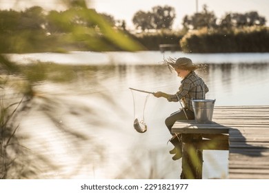 a cute blond boy in a straw hat, jeans and rubber boots with a fishing rod on a wooden pier is fishing on a sunny, warm day - Powered by Shutterstock