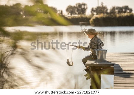 a cute blond boy in a straw hat, jeans and rubber boots with a fishing rod on a wooden pier is fishing on a sunny, warm day