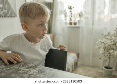 A cute blond boy-child is sitting at a table in the kitchen and watching TV. Fun leisure while waiting. Side view