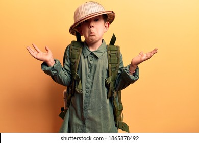 Cute Blond Kid Wearing Explorer Hat And Backpack Clueless And Confused With Open Arms, No Idea And Doubtful Face. 