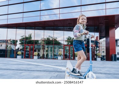 Cute blond little Caucasian girl wear patchwork cardigan and jeans shorts enjoy having fun riding scooter city street outdoors sunny day. Healthy sport children activity outside - Powered by Shutterstock