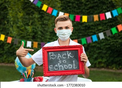 Cute Blond Teen Boy In Medical Mask Posing With Small School Board Against Flags Background.