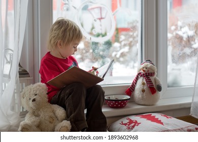 Cute Blond Toddler Child, Sitting On The Window, Looking At The Snow Falling Outside, Reading A Book