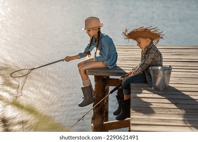 cute blonde boy and girl in straw hats and rubber boots with a fishing rod and a scoop on a wooden pier fishing on a sunny, warm day - Powered by Shutterstock