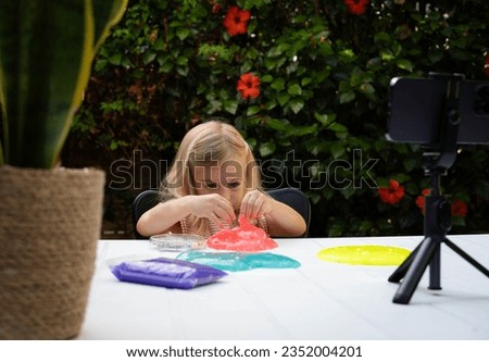 Cute blonde caucasian baby girl playing with slime and watching video lesson or cartoon on smart phone. Online education, classes at home, on distance. Child sensory development game.