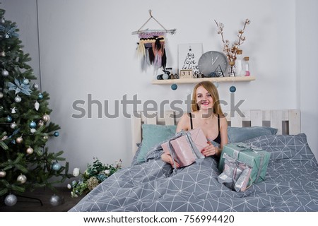 Cute blonde girl on black bra lying on bed with gifts boxes against new year tree at studio. Happy holidays.
