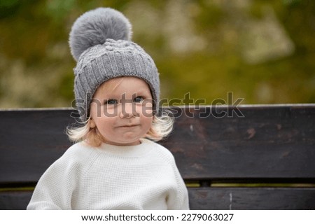 Cute blonde kid sitting on a bench outside in a park, smiling. Happy child wearing a hat outdoors on a spring day. Adorable grinning caucasian boy resting on a bench after play time on a playground.