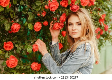 A Cute Blonde Stands Near A Bush With Red Roses. Attractive European Woman In A Denim Jacket.