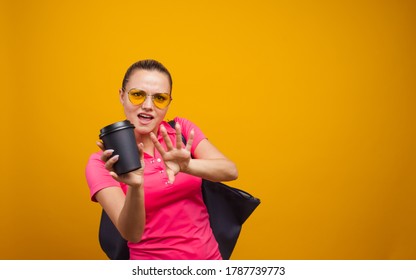 Cute blonde in sunglasses and t-shirt with bag on shoulder, drinking her hot drink, portrait on yellow background copy space