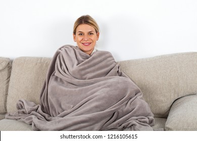 Cute blonde woman relaxing o the sofa wrapped in a soft grey blanket, white wall in the background