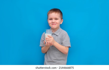 Cute Boy 5-6 Years Old Drinking Milk On Colored Blue Background. He Knows That He Needs To Drink Milk For Bone Health. The Baby Loves Milk. Concept Of Children's Healthy Nutrition.