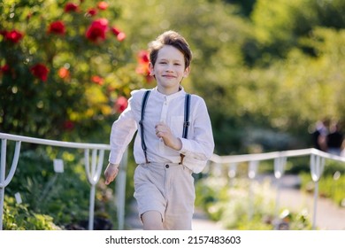 A cute boy of 7 years old in a shirt walks in the garden. Sincere smile of a child. Classic clothing style men's white shirt and suspenders. Summer portrait of a running kid retro style. carefree 