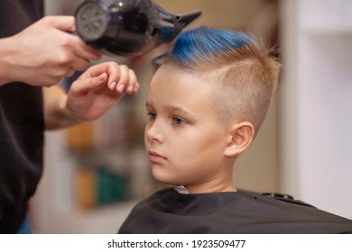 A Cute Boy In A Beauty Salon At A Hairdresser, A Stylist Dries A Child's Blue Dyed Hair, A Barbershop Job, A Fashionable Hairstyle For A Schoolboy.
