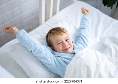 a cute boy in a blue bathrobe wakes up at home in the morning in a bed with white linens, rubs his eyes, yawns, smiles, stretches after sleep, a healthy sleep for children - Powered by Shutterstock