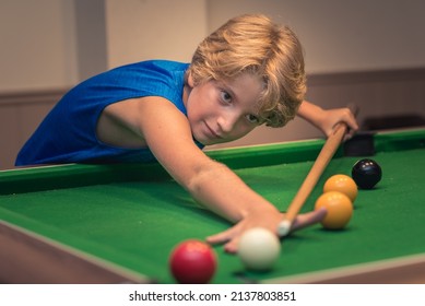 Cute boy in blue t shirt plays billiard or pool in club. Young Kid learns to play snooker. Boy with billiard cue strikes the ball on table. Active Leisure, sport, hobby concept

