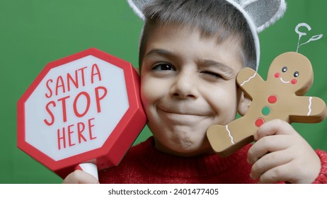 Cute boy with christmas sweater and deer alntlers holding sign Santa stop here and gingerbread man. Isolated on green background,. High quality photo - Powered by Shutterstock