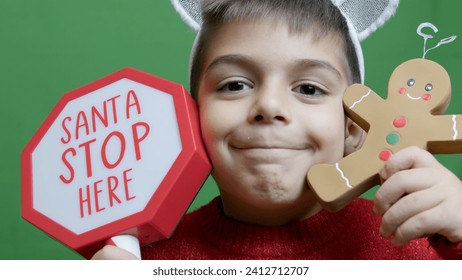 Cute boy with christmas sweater and deer alntlers holding sign Santa stop here and gingerbread man. Isolated on green background,. High quality photo - Powered by Shutterstock
