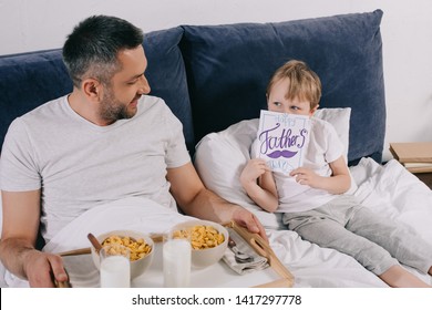 Cute Boy Covering Face With Fathers Day Greeting Card While Sitting In Bed Near Daddy With Breakfast On Tray