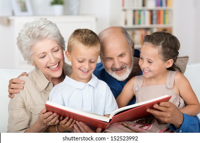 Cute Boy And Girl Sitting In The Lap Of Their Grandparents And Looking Happily Together At A Photo Album