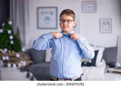 A cute boy in glasses, an elegant shirt straightens his bow tie in a white room. A handsome boy, well dressed for Christmas. A young handsome guy, smiling. an emotional and cheerful guy