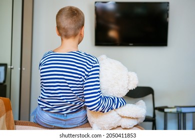 Cute Boy And His Plush Toy Bear Are Watching Tv While Sitting On The Couch In The Living Room At Home. View From The Back. Young Guy In Striped T-shirt And Blue Jeans