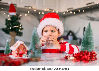 Cute Boy Kid In A Red Santa Hat Drinking Filtered Water From A Glass In The Kitchen. Holidays, Health Concept.